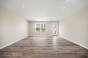 Empty room with light-colored walls, a large window, and wooden flooring. A door is situated near the window.