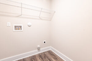 Empty laundry room with a white wire shelf along the upper wall, utility connections for a washing machine, and a dryer vent on the lower wall. The floor is wood laminate.