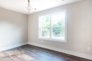 An empty room in the serene Poplar Village, with light beige walls, a large window, and dark wood flooring that exudes elegance. A ceiling light fixture is centered above, casting a warm glow.