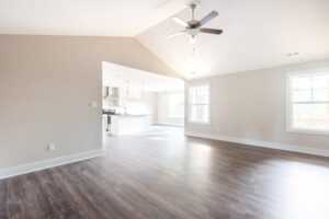This spacious room in Poplar Village features light wood flooring and large windows that offer a view into a modern kitchen. A ceiling fan hangs from the vaulted ceiling, adding elegance to this Belvedere-inspired space.