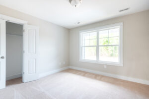 A small, empty, carpeted room with beige walls, a double door closet, and a large window with white trim letting in natural light.