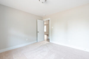 A vacant room with beige carpet, light-colored walls, a closed window, and an open door leading to another room. A ceiling light fixture is centered above.