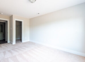 A bright, empty room with light beige carpet, neutral-colored walls, and white trim. The room has a door leading to a closet and another to a hallway.