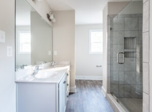 A modern bathroom with a dual-sink vanity, large mirror, walk-in glass shower, and wood-look flooring. Natural light enters through two windows.