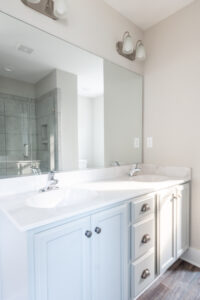 A modern bathroom with a large mirror, double sink vanity, light gray cabinetry, overhead light fixtures, and a glass-enclosed shower in the background.