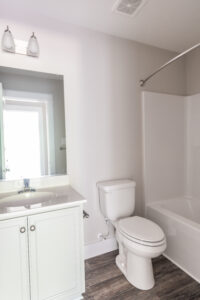 A bathroom with a white sink and vanity, a toilet, and a bathtub shower combination. The bathroom has a large mirror and wood-patterned flooring.