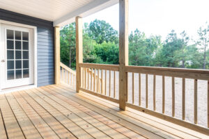 The wooden deck with railing, a charming feature of the house with blue siding in Poplar Village, offers a warm welcome through its white door with glass panels. In the backdrop, trees create a serene setting for this Belvedere-inspired retreat.