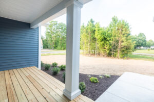 From a charming wooden porch with blue siding in Poplar Village, enjoy a belvedere view of a garden bed dotted with small plants. Beyond, a patch of dirt flows into lush grass and trees that whisper tales of nature's beauty.