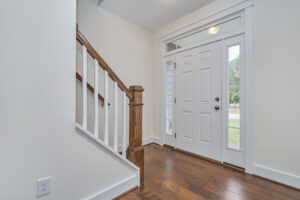 An entryway with wooden stairs, white balusters, hardwood floor, and a white front door with a sidelight and transom window.