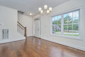 An empty room with hardwood floors, white walls, a large window, a wooden staircase with a white banister, and a chandelier hanging from the ceiling.