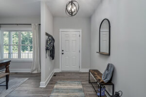A modern entryway with a white door, a round mirror above a wooden bench, and coats hanging on wall hooks. There is a window with white curtains on the left and a rug on the floor.