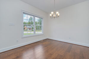 An empty room with light-colored walls, a window facing outside, a hanging light fixture, and brown wooden flooring.