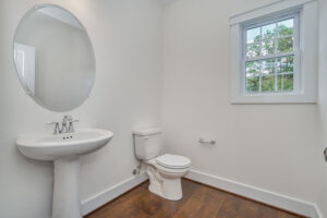A clean, minimalist bathroom with a pedestal sink, round mirror, and a toilet on wooden flooring. A small window allows natural light to enter.