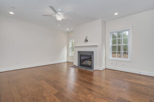 A spacious living room with hardwood floors, a ceiling fan, a large window, and a fireplace centered between two smaller windows. The walls and ceiling are painted white.
