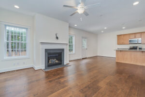A spacious, unfurnished living area with hardwood floors, a ceiling fan, a fireplace, and a modern kitchen visible in the background. Large windows allow natural light into the room.