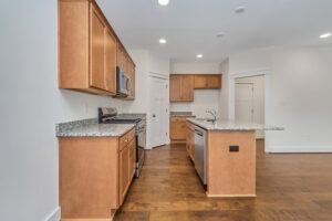 A modern kitchen with wooden cabinets, granite countertops, stainless steel appliances, a central island with a sink, and hardwood floors.