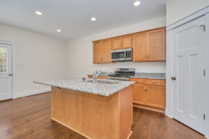 A modern kitchen with light wood cabinets, granite countertops, an island, stainless steel appliances, and recessed lighting. A door and white door on the right lead to other areas.