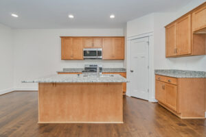 A modern kitchen with light wooden cabinets, granite countertops, an island, stainless steel appliances, and a hardwood floor.