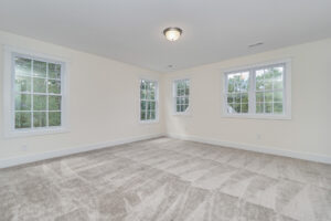 A vacant room with light beige walls, three large windows with white trim, and a neutral-toned, carpeted floor. Ceiling light fixture centered.