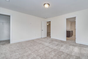 A carpeted bedroom with white walls, a ceiling light, open door to a walk-in closet, and another open door to a bathroom with double sinks.