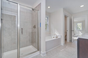 A bathroom with a glass-enclosed shower, a white bathtub, tiled floors, beige walls, a window, and a view of another room through the open door.
