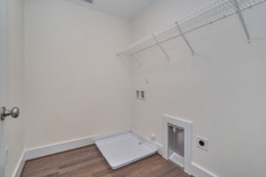 A laundry room with white walls, a wall-mounted wire shelf, and hookups for a washer and dryer. The floor is brown wood, and there's a square white tray area for the washer.