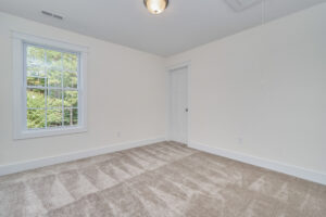 Empty, carpeted room with off-white walls, a window showing green trees outside, white trim, a flush-mounted ceiling light, a door, and a ceiling hatch with a pull string.