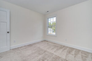 A small empty room with beige carpet, white walls, a white door, and a single window showing an outside view of houses and trees.