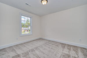 A vacant room with light beige walls, a single window showing an outside view, a ceiling light fixture, and a carpeted floor.