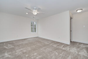 A spacious room with beige carpet, white walls, a ceiling fan with light, a single window, and a door leading to another area.