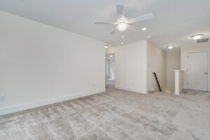 A carpeted room with a ceiling fan, recessed lighting, and light-colored walls. A hallway with a staircase and doors is visible in the background.