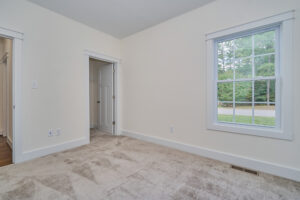 A small empty room with beige carpet, a large window on the right, and a doorway on the left leading to a closet or another room. The walls are painted a light color.