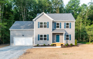 Nestled in the serene Poplar Village, this two-story gray house with blue shutters features a charming front porch and an attached two-car garage, all set against a lush, wooded backdrop.