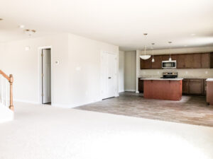 A modern open-concept living space with a kitchen featuring wooden cabinets, an island, and stainless steel appliances. The living area has light carpeting and a wooden staircase railing.