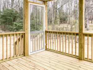 A wooden deck with a screened door opens to a wooded area. The deck has simple vertical railings and a natural wood finish. Trees and vegetation are visible in the background.