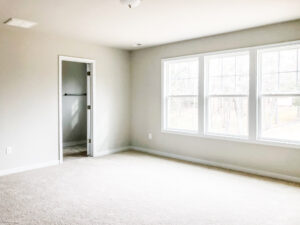 A bright, empty room with carpeted floor, neutral-colored walls, three large windows, and an open door leading to another space.