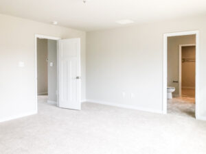 A carpeted empty room with white walls, two doors, and a doorway to a bathroom with a visible toilet.