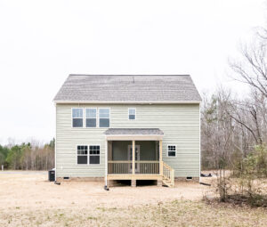 A two-story house with light green siding, multiple windows, and a small covered porch. The surrounding yard is mostly bare with some trees visible in the background.