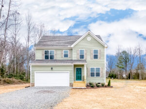A two-story house with light green siding, a dark gray roof, a double garage, and a green front door. The yard is barren, and trees are in the background under a partly cloudy sky.