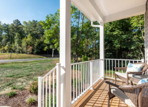 A white porch with a wood deck, two wicker chairs, and a small table overlooks a grassy area and trees under a clear blue sky.