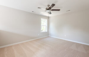 An empty room with beige carpet, light gray walls, a ceiling fan with light, and a single window allowing natural light in.