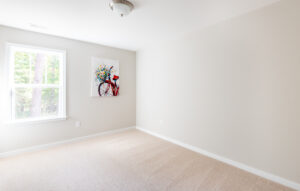 An empty room with beige carpet, white walls, a window on the left side, and a painting of a red bicycle with flowers hanging on the wall.