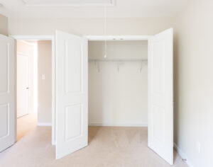 An empty closet with double doors, beige walls, and a beige carpet. The closet has a metal rod for hanging clothes.