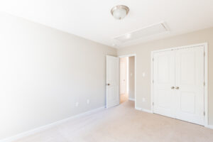 An empty room with beige carpet, white walls, a two-door closet, an open door leading to a hallway, and a ceiling light fixture.