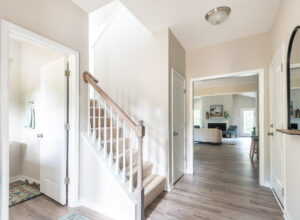 A home interior showing a carpeted staircase with a wooden handrail, doors to the left and right, and an open view into a living area with light-colored walls and wood flooring.
