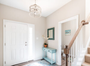 A bright entryway with a white door, light beige walls, a turquoise console table with a potted orchid, a mirror above it, a stairway to the right, and a pendant light overhead.