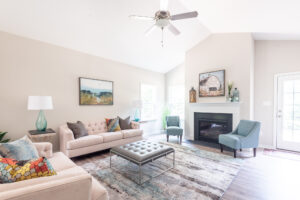 A bright, modern living room with two beige sofas, two blue armchairs, a central ottoman, a fireplace, and a ceiling fan. Art and decor items are placed throughout the space.