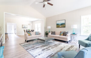A bright, modern living room features two beige sofas, a glass coffee table, a patterned rug, and turquoise armchairs. Lamps and art adorn the space. In the background is a connected dining area.