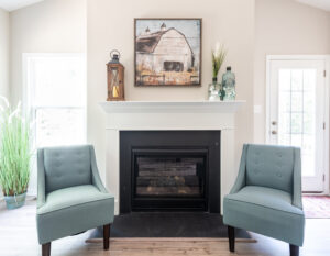 A living room with a white fireplace, two light blue chairs, a framed picture above, potted plants on the hearth, and a glass-paneled door on the right.