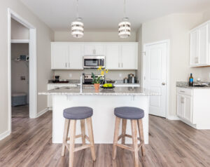 A modern kitchen with a central island featuring two stools, granite countertops, a microwave, and white cabinetry. Hanging pendant lights illuminate the space.